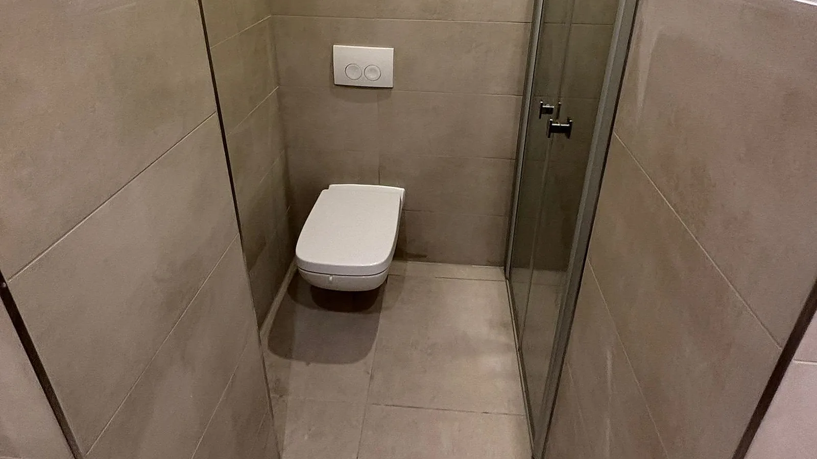 Wand-WC am Ende eines schmalen Raums, seitlich eine Glastür