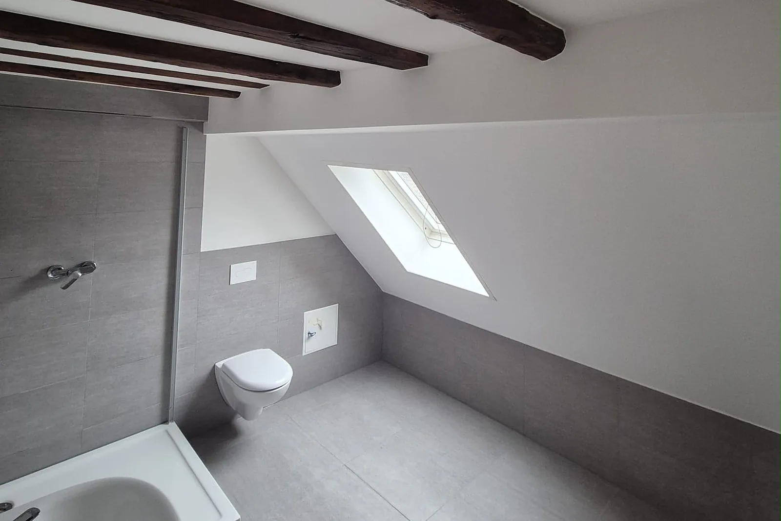 Dachgeschossbad mit Dachfenster, dunklen Holzbalken und grauen Wandfliesen