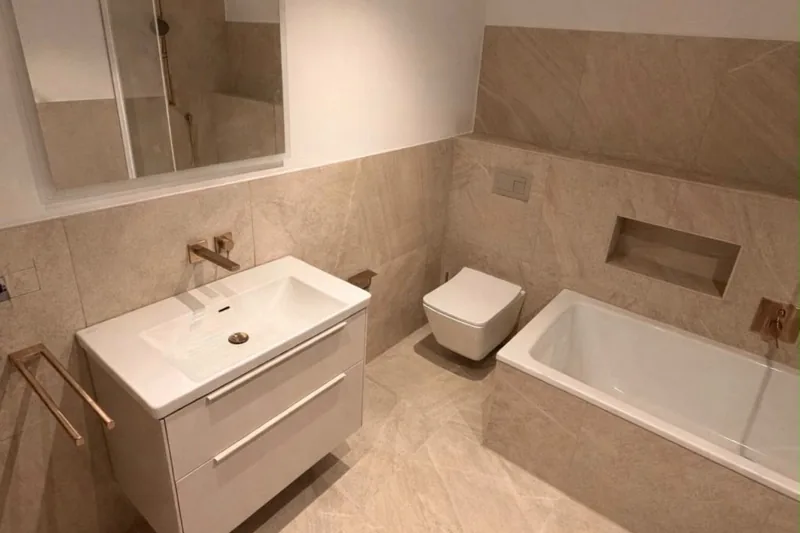 Badfliesen: Waschtisch mit Unterschrank vor großen Fliesen in Beige, dahinter Wand-WC und Badewanne