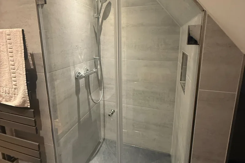 Duschabtrennung: Glasduschtür mit verchromten Scharnieren vor einer Dusche mit grauen Großformatfliesen